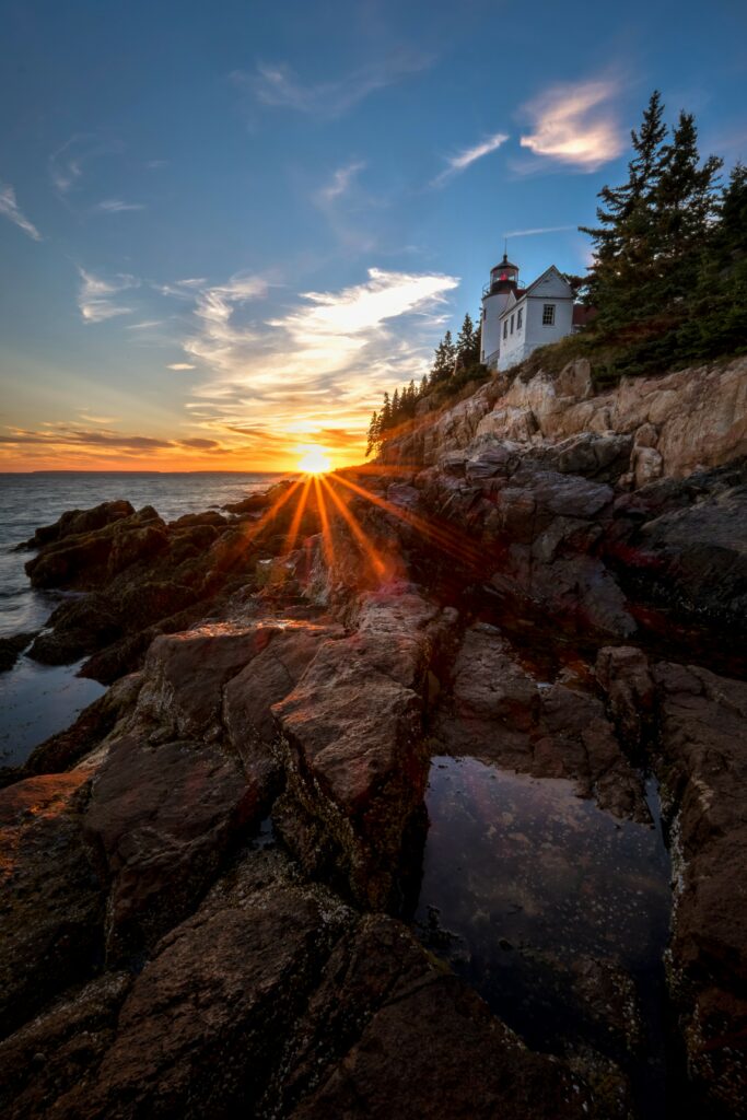 Classic lighthouse on rugged rocks with sun rays and coastal buildings at sunset wallpaper background