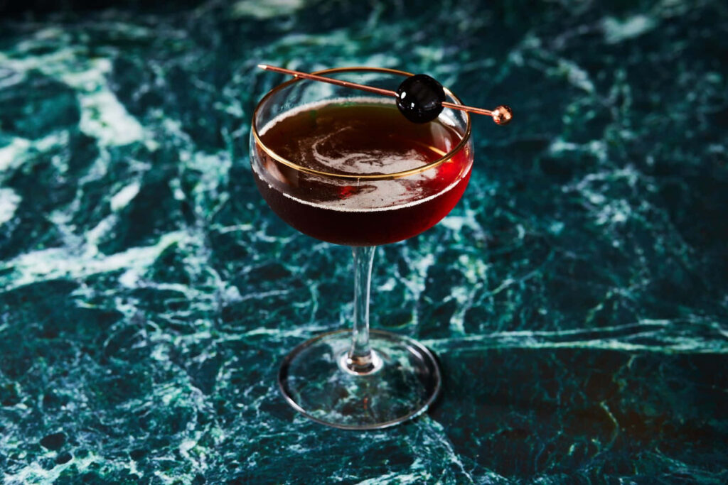 Elegant Manhattan cocktail in coupe glass with cherry on deep green marble wallpaper background