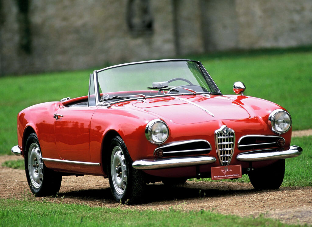 Classic red Alfa Romeo Spider convertible parked on grassy path with vintage stone wall background Wallpaper