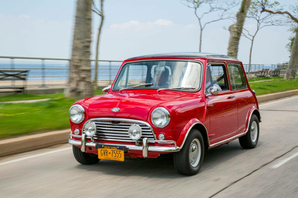 Red classic mini car driving on scenic tree-lined road vintage nostalgic wallpaper background