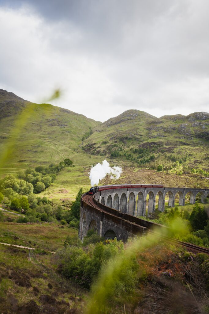 Classic steam train crossing curved stone bridge with white smoke and green hills background Wallpaper