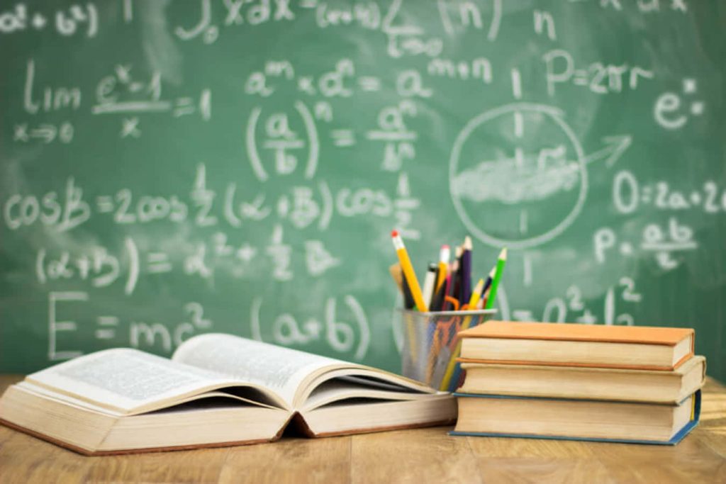 Open book and stacked textbooks with writing tools on table and chalkboard formulas background Wallpaper