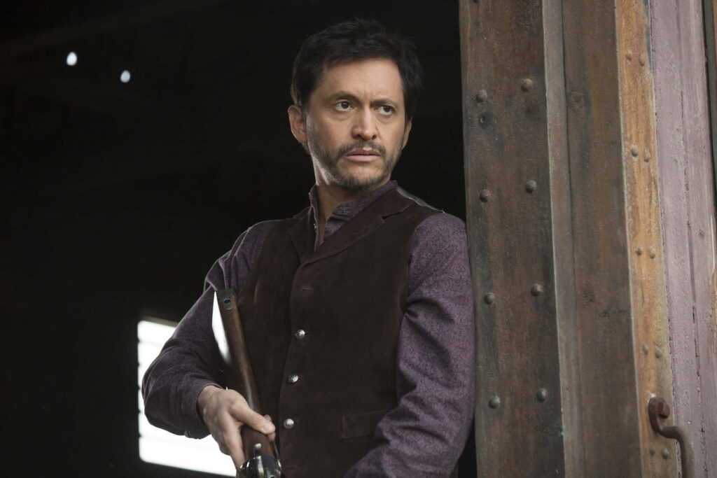 Clifton Collins Jr in rustic doorway with rifle wearing dark vest gritty Western suspense wallpaper background