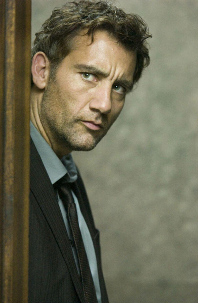 Clive Owen in dark suit gazes thoughtfully behind wall with muted wallpaper cinematic mood