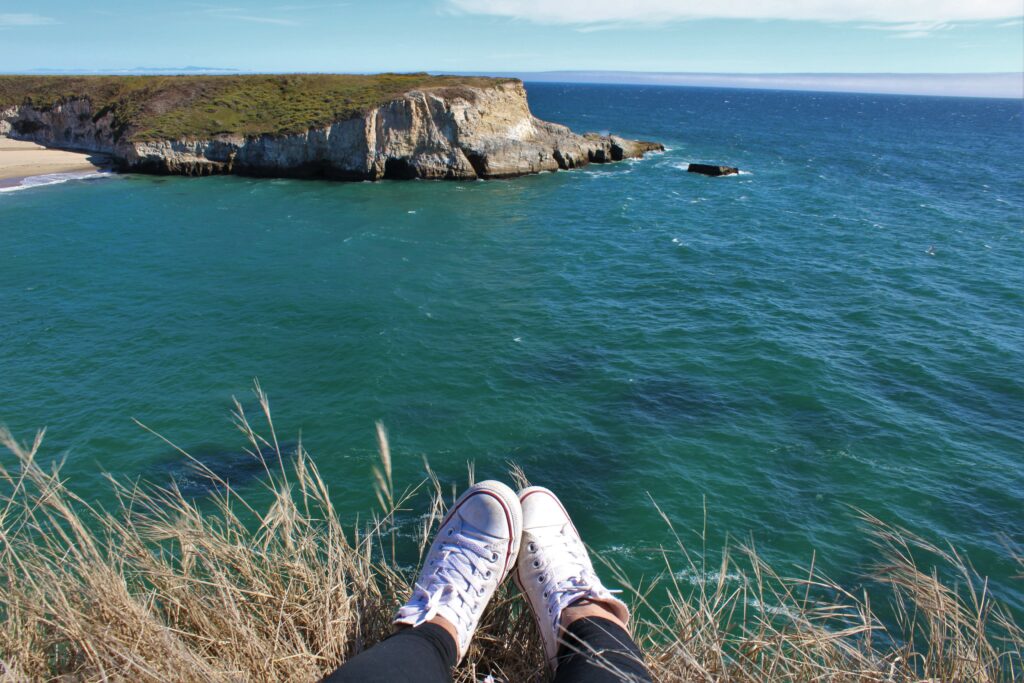 White sneakers on grassy cliffs overlooking turquoise sea and rugged coastline under blue sky Wallpaper