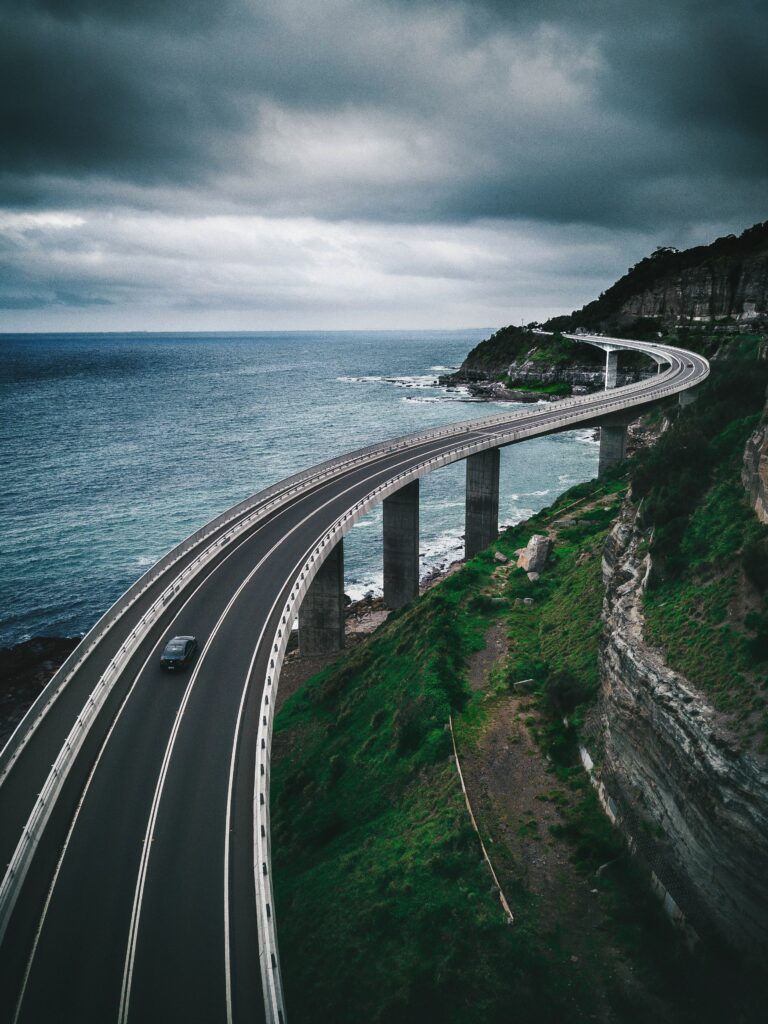Sweeping coastal bridge curves above turquoise sea and rugged mountains under dramatic skies Wallpaper