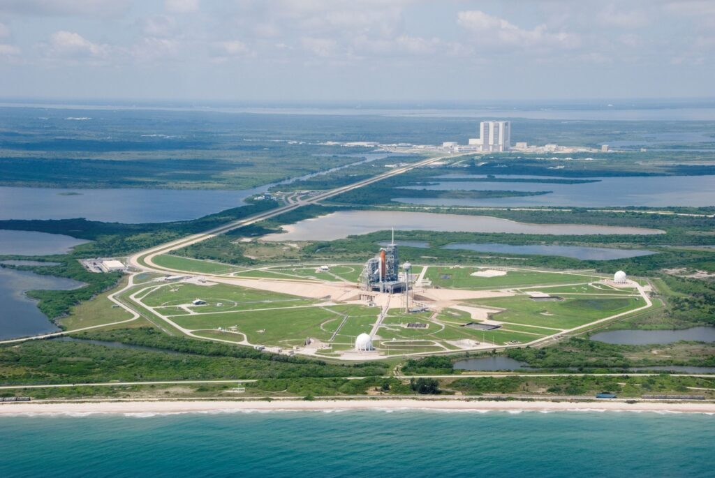 Space shuttle launch pad on coastal complex surrounded by water and green land under clouds Wallpaper