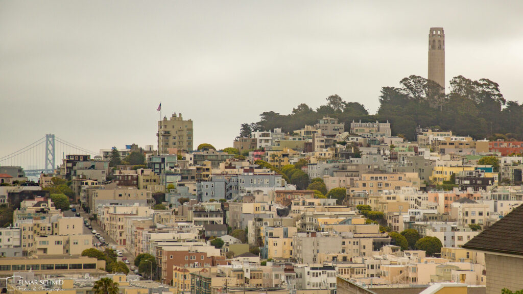 Coit Tower over colorful San Francisco buildings with Bay Bridge and cloudy sky background Wallpaper