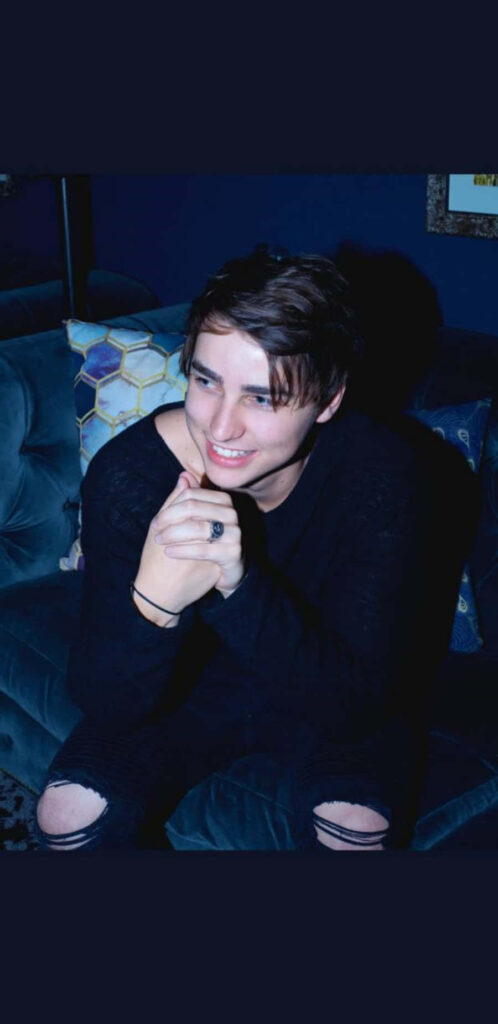 Colby Brock smiling on blue sofa wearing black sweater and ripped jeans with geometric pillows Wallpaper