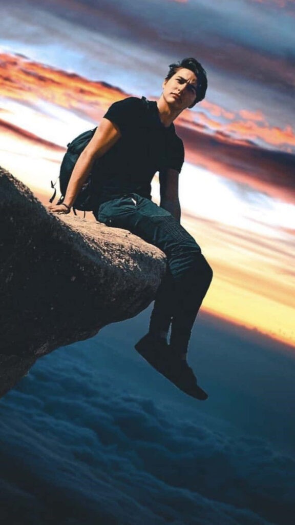 Colby Brock sitting on rocky cliff at vibrant sunset with colorful sky clouds adventure scene Wallpaper