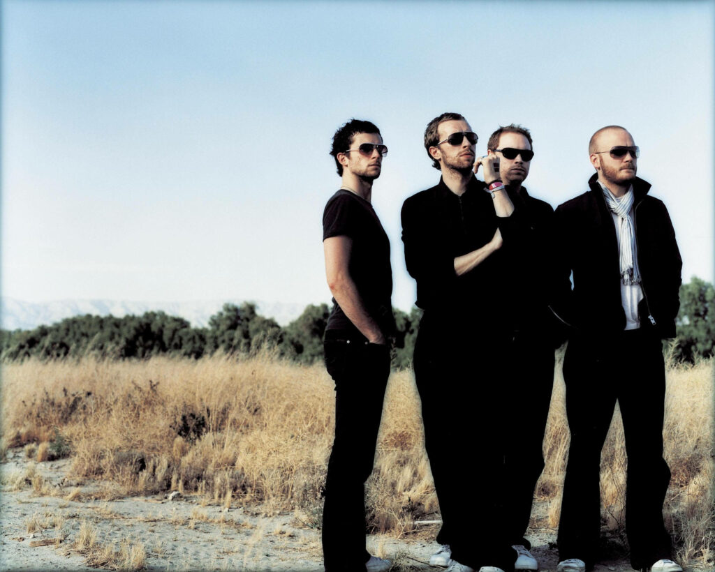 Coldplay band group photo in sunlit meadow with dark clothes sunglasses scenic nature background Wallpaper