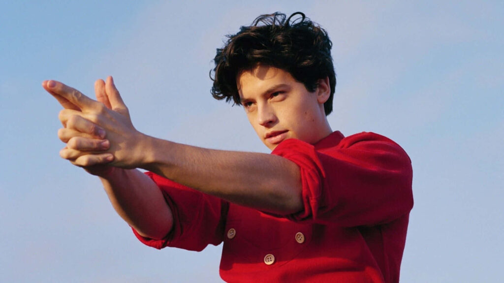 Cole Sprouse in vibrant red jacket outdoors striking dramatic pose against clear blue sky wallpaper