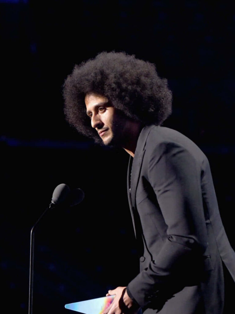 Colin Kaepernick in black suit at microphone with iconic afro and focused expression wallpaper