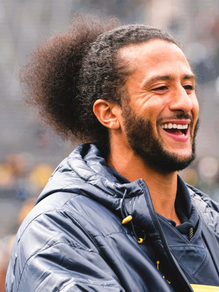 Colin Kaepernick mid-laugh in navy jacket vibrant wallpaper with blurred stadium background