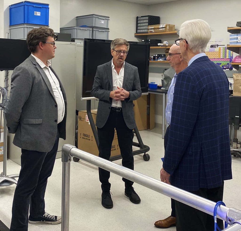 Men in business-casual discussing in a modern well-lit lab with equipment and storage units background Wallpaper