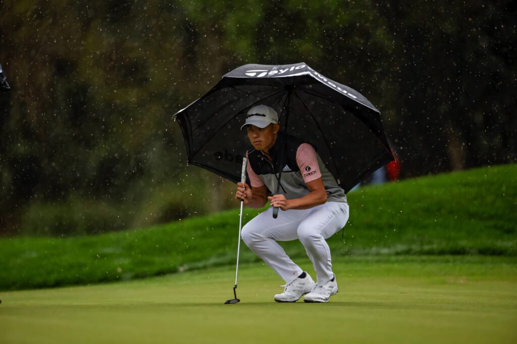 Collin Morikawa focused on rainy golf course holding black umbrella in white and grey sportswear Wallpaper