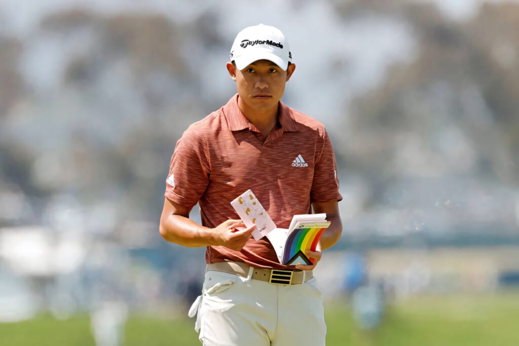 Collin Morikawa in rust Adidas polo on golf course holding notebook focused on next golf shot Wallpaper