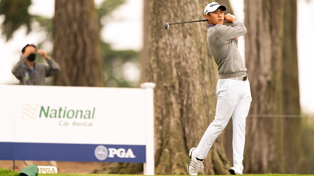 Collin Morikawa mid-swing at PGA League on lush green golf course with tall trees background Wallpaper