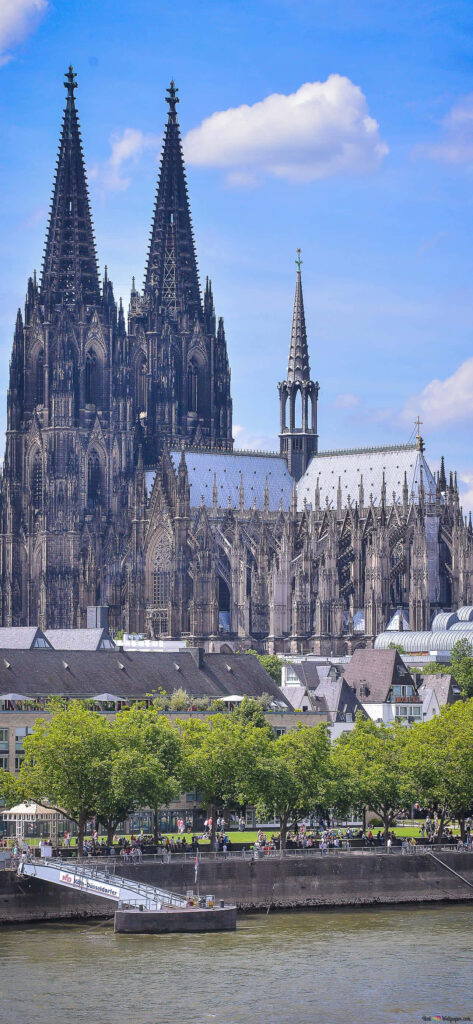 Cologne Cathedral Gothic spires and stonework with riverside buildings and blue sky background Wallpaper
