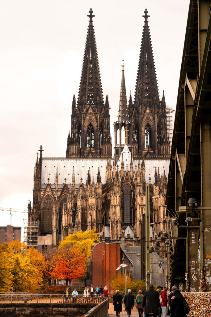 Cologne Cathedral in autumn with golden leaves and people walking along riverside path wallpaper