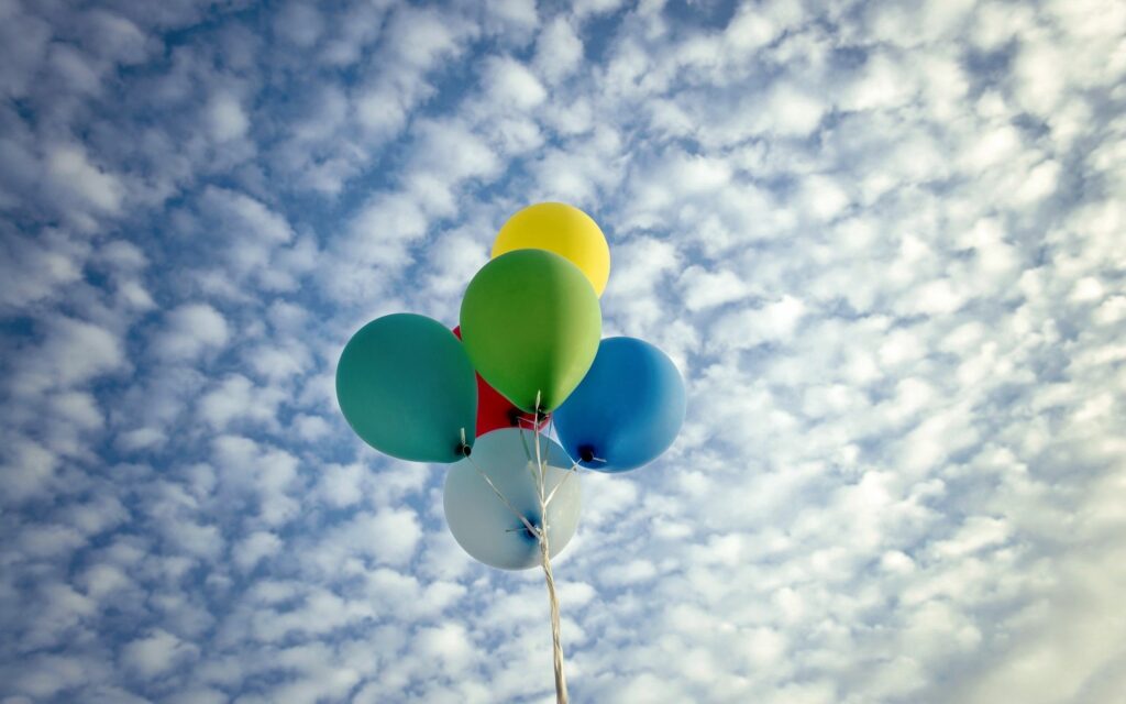 Colorful balloons rising against bright blue sky with fluffy clouds creating cheerful wallpaper