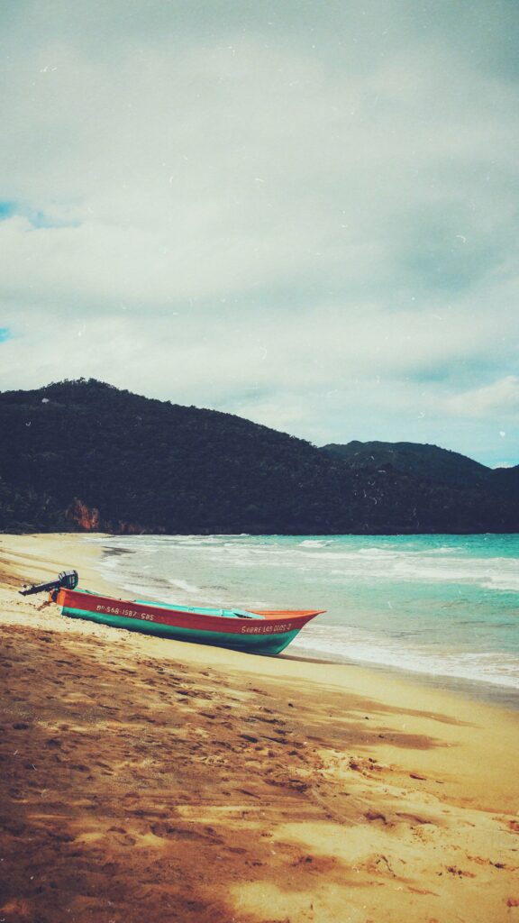 Colorful boat on sandy beach with turquoise sea waves and lush mountains under cloudy sky Wallpaper