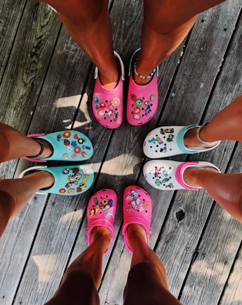 Friends wearing colorful Crocs with playful charms standing in a circle on wooden deck summer Wallpaper