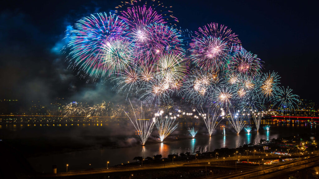 Colorful fireworks illuminate night city skyline and reflect on water creating festive atmosphere Wallpaper