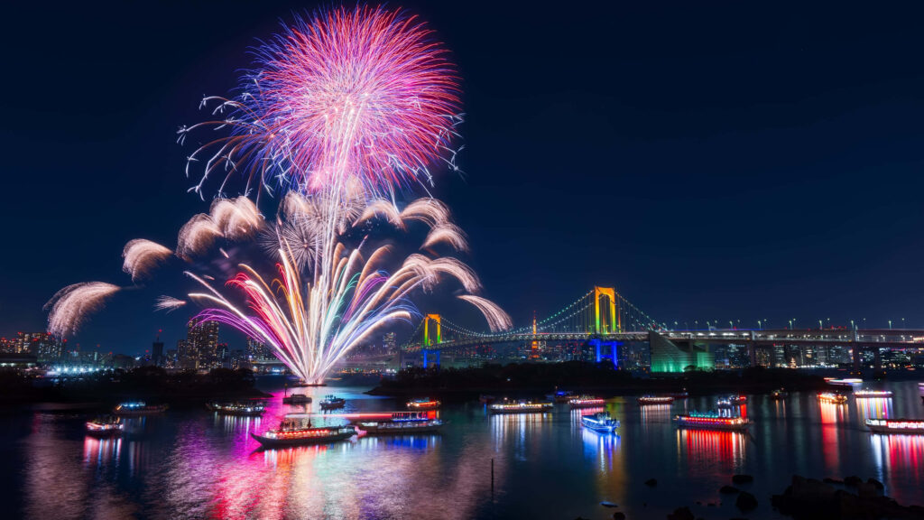 Colorful fireworks illuminate city skyline and water with boats and glowing bridge at night Wallpaper