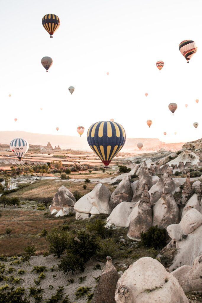 Colorful hot air balloons float above rugged rock formations and mountains in soft natural light Wallpaper