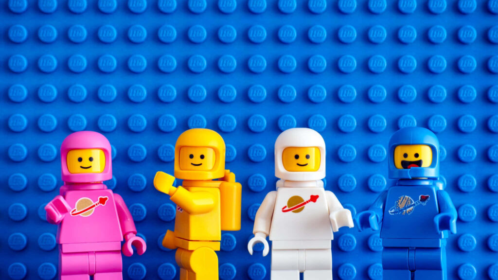 Four colorful Lego astronaut minifigures in pink yellow white and blue on blue Lego background Wallpaper