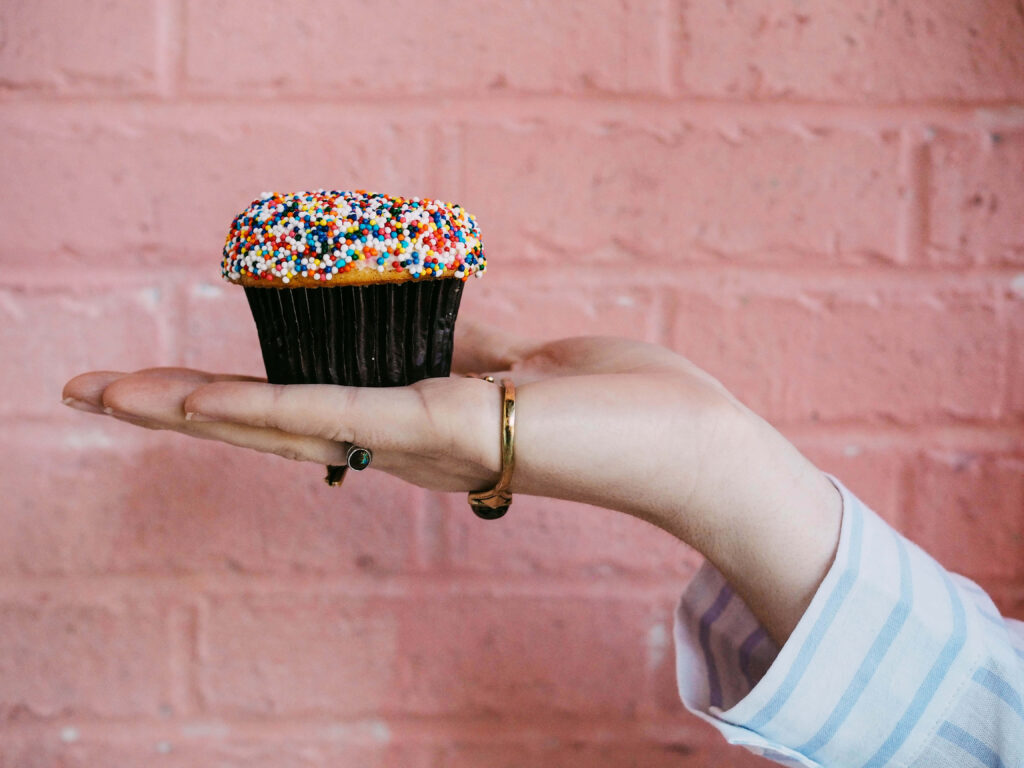 Hand holding a cupcake with rainbow sprinkles against pastel pink brick wall background Wallpaper