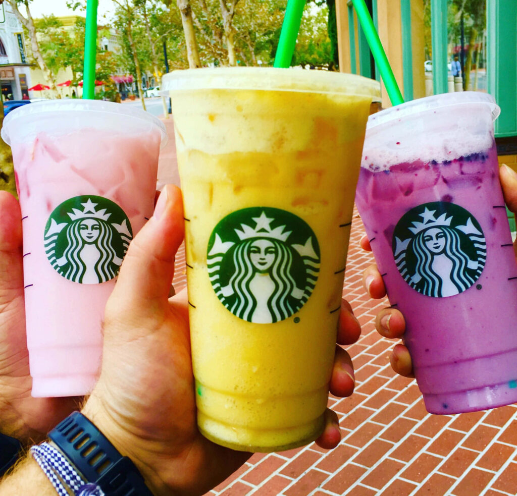 Three hands hold colorful Starbucks iced drinks pink yellow and purple outside Starbucks store Wallpaper