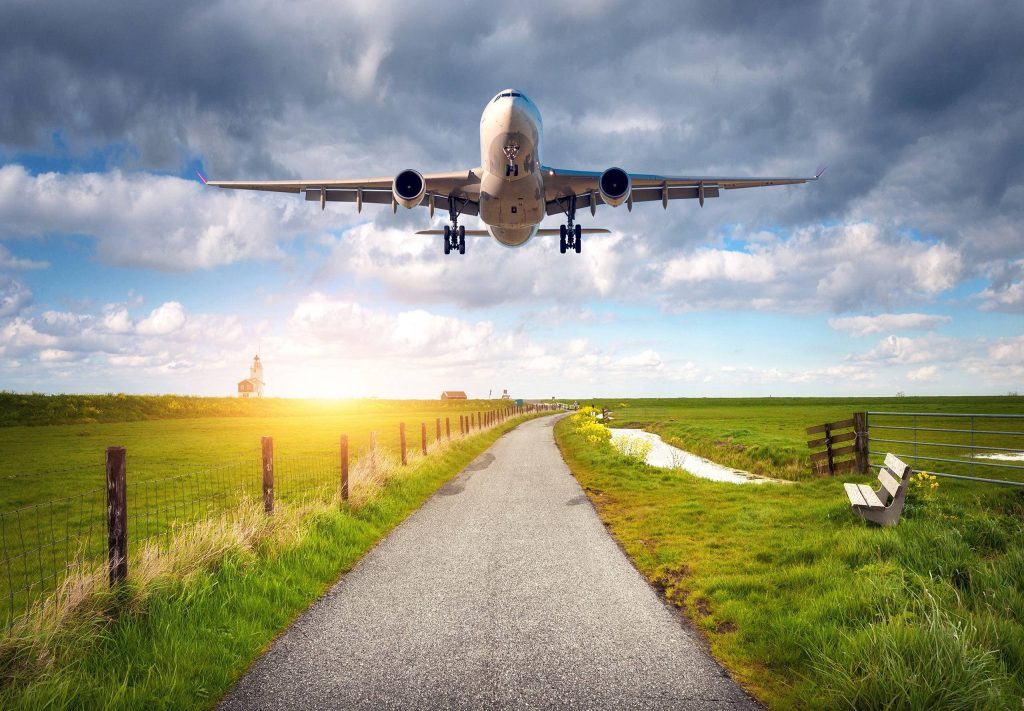 Commercial airplane flying over green countryside with winding road and lighthouse at sunset Wallpaper