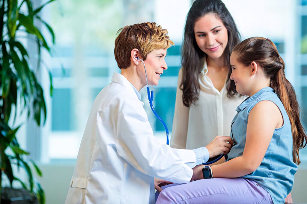 Doctor checking young girl’s heartbeat with stethoscope while mother smiles in bright clinic Wallpaper