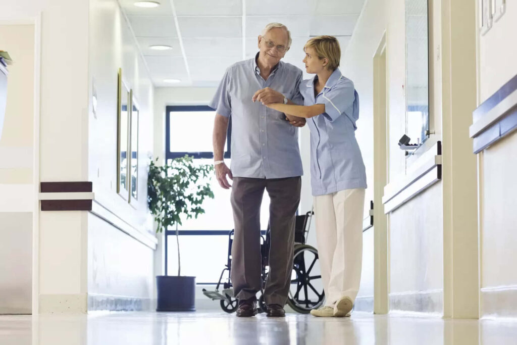 Nurse helping elderly man walk in sunny hospital corridor near wheelchair showing compassionate care Wallpaper