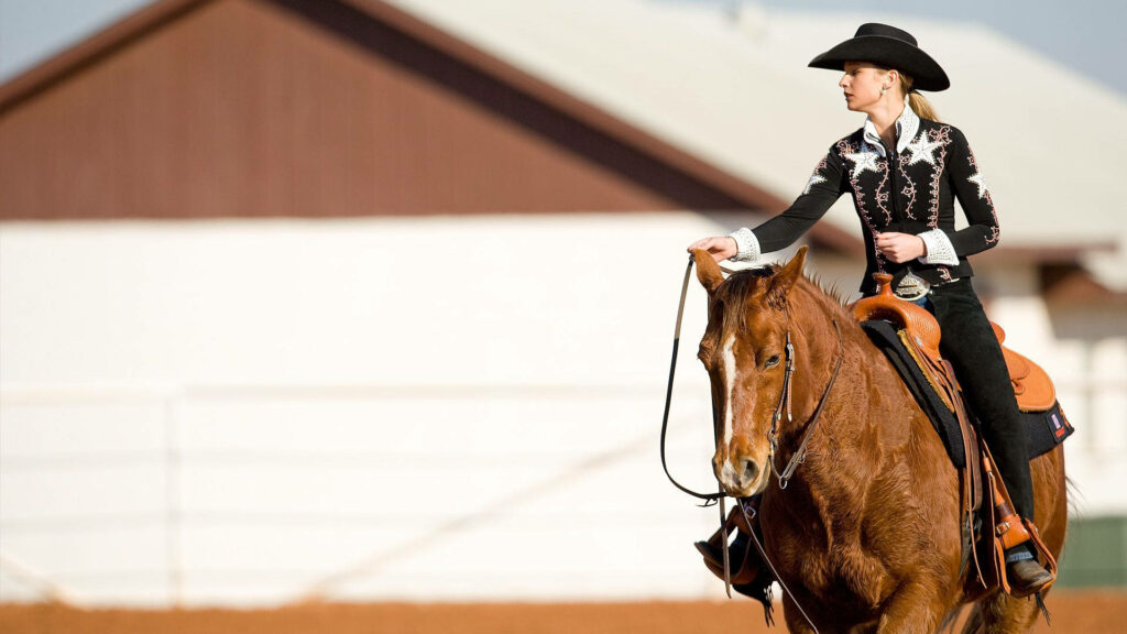 Cowgirl in black hat riding brown horse in reddish dirt arena near sunlit barn western scene Wallpaper