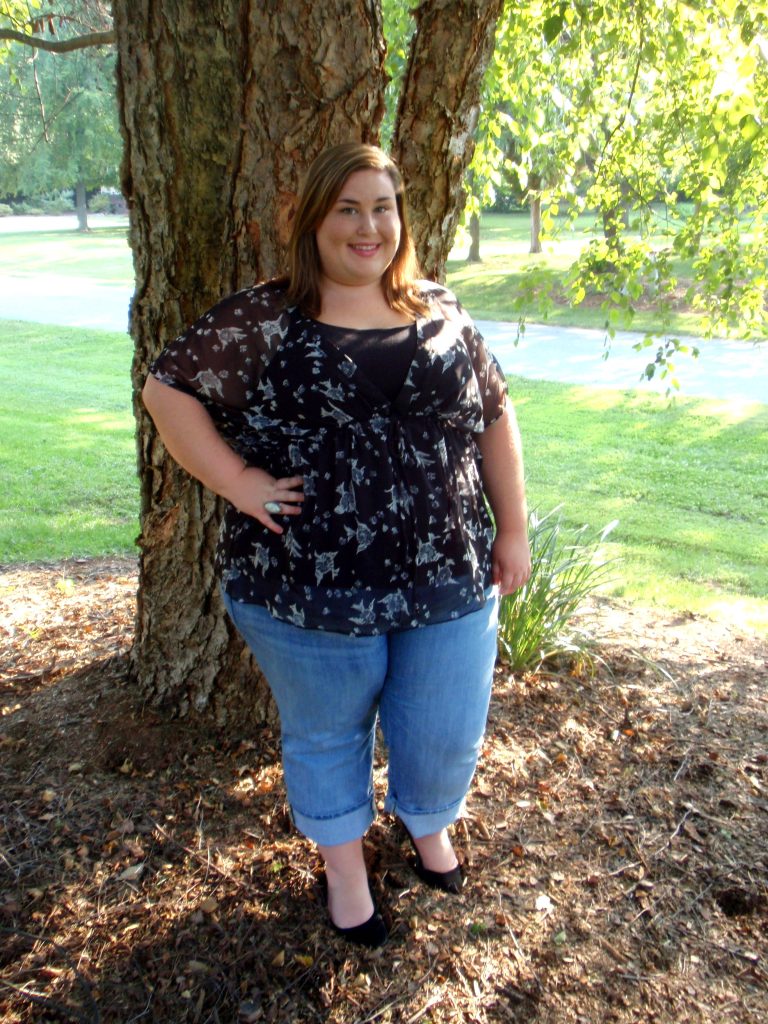 Plus-size woman in black floral sheer top and jeans joyfully posing beside tree outdoors Wallpaper
