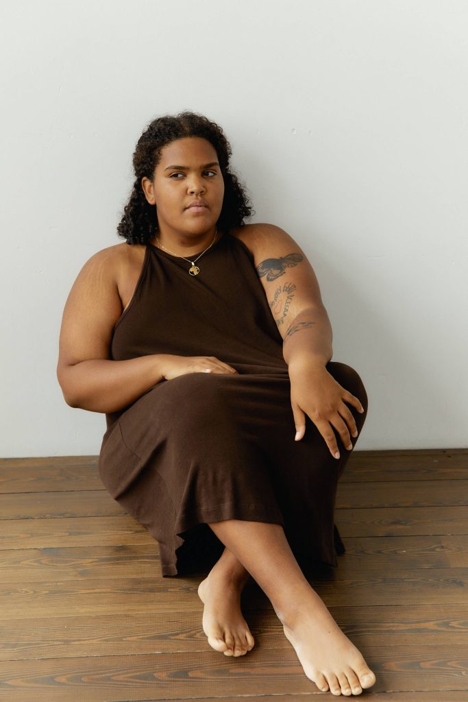 Plus size woman in brown dress seated on wooden floor with natural lighting and white wall background Wallpaper