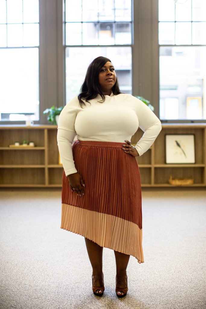 Confident plus size woman in cream ribbed top and pleated skirt posing in modern office space Wallpaper