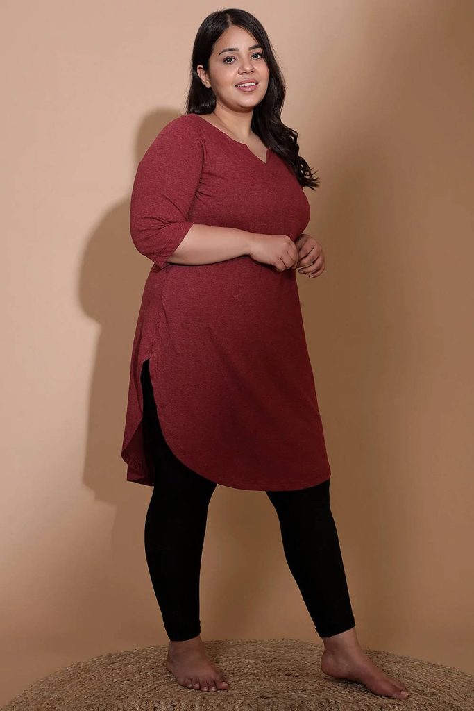 Plus size woman in dark red tunic and black leggings stands barefoot on woven mat studio portrait Wallpaper