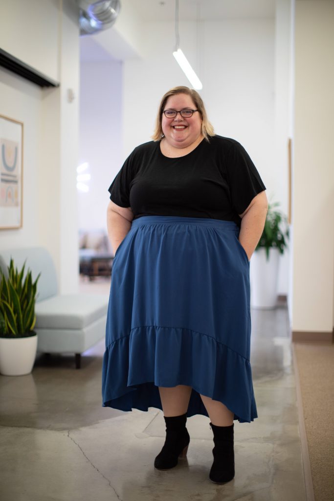 Plus-size confident woman smiling in modern office wearing black top blue skirt and ankle boots Wallpaper