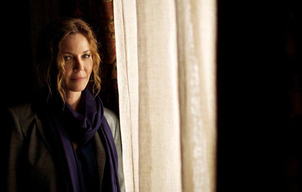 Connie Nielsen in warm natural light with wavy hair and elegant scarf by beige curtains Wallpaper