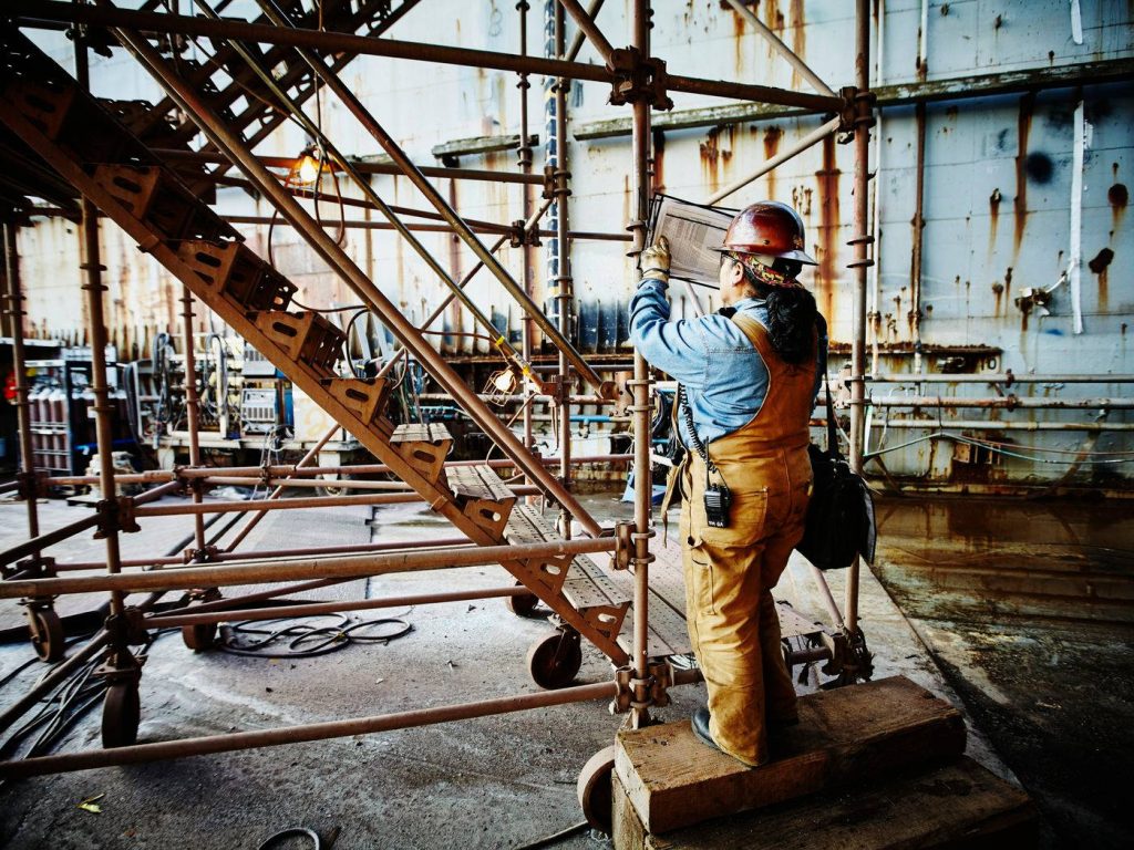 Construction worker in hard hat reviewing blueprints on scaffolding with rusted metal backdrop Wallpaper