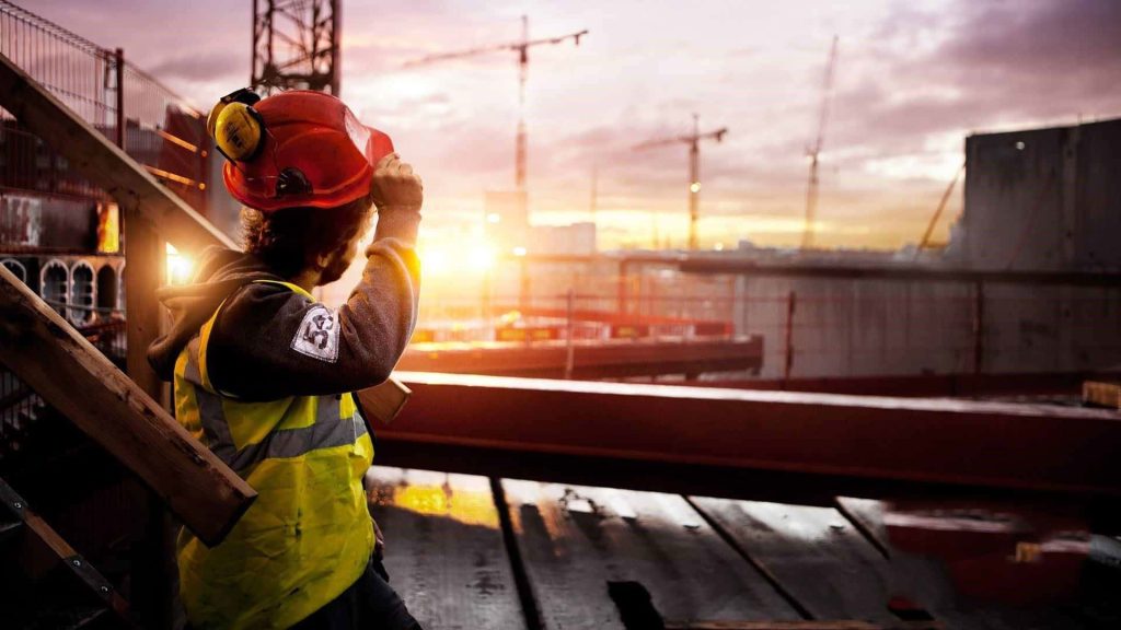 Construction worker in red hard hat and vest at building site with cranes at sunset background Wallpaper