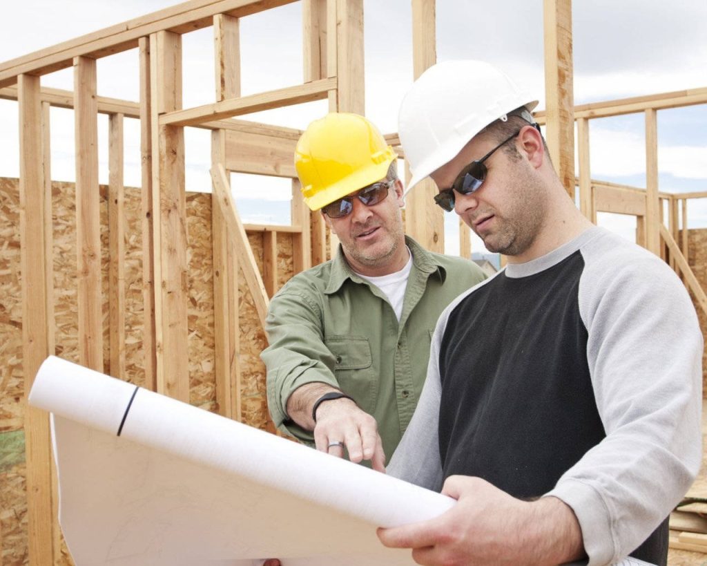 Two construction workers in hard hats study blueprints by wooden framing on bright site Wallpaper