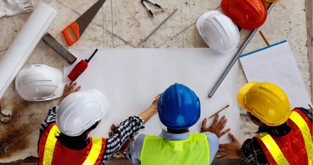 Top-down view of construction workers in safety vests and helmets reviewing blueprint at site Wallpaper