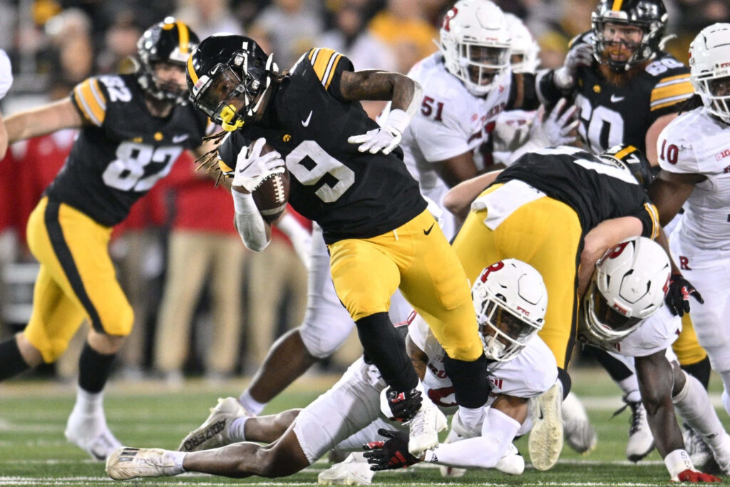 Cooper DeJean in black and gold Iowa Hawkeyes uniform dodging defenders on green football field Wallpaper