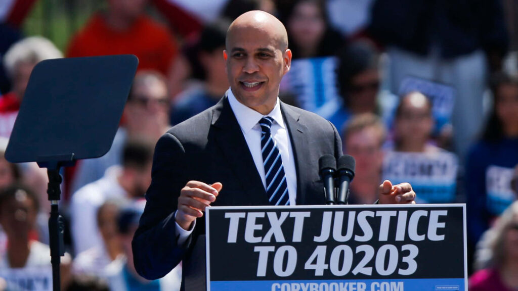 Cory Booker speaking at rally with justice campaign sign vibrant outdoor background energy Wallpaper