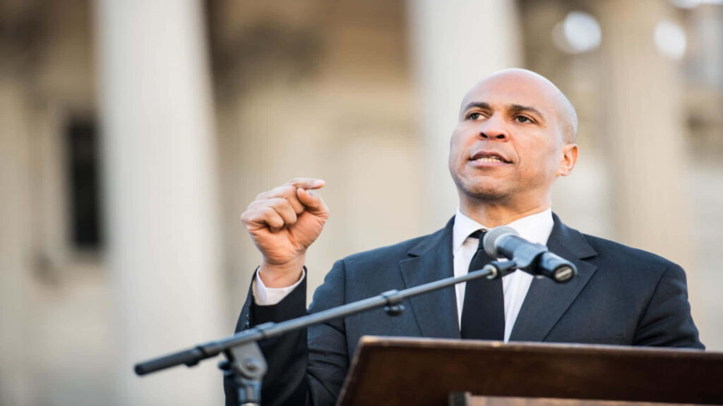 Cory Booker delivering passionate speech in suit behind podium with historic columns background Wallpaper